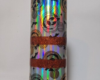 May include: A tall, silver tumbler with a Halloween theme. The tumbler features a pattern of jack-o'-lanterns and bats, with bands of orange glitter. The design has a holographic effect, creating a rainbow sheen.