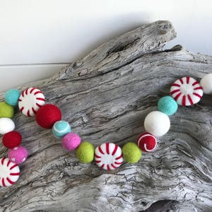 Puede incluir: Una guirnalda festiva hecha de bolas de fieltro y formas de caramelo de menta en colores rojo, blanco, verde, rosa y azul. La guirnalda está drapeada sobre un trozo de madera a la deriva.