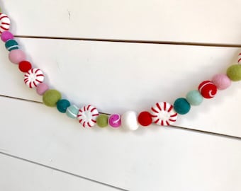 May include: A festive garland made of felt balls in various colors, including pink, green, red, and white. The garland features peppermint candy designs and is perfect for decorating for the holidays.