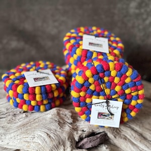 Könnte beinhalten: Set mit vier bunten Filz-Untersetzern in einem blau-rot-gelben Farbschema. Die Untersetzer bestehen aus Wollfilzkugeln und sind handgefertigt. Die Untersetzer sind mit einem kleinen Etikett verpackt, auf dem "Wooly Sheep 100% Wool Felt Creations" steht.