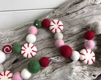 May include: A festive garland made of felt balls in pink, white, red, and green. The garland features peppermint candy designs and a swirl design.