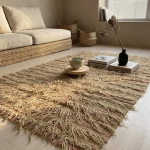 May include: A beige, shaggy rug with a textured appearance, placed on a light-colored wooden floor. The rug is rectangular and appears to be made of natural fibers. A sofa, baskets, books, and a vase are also visible in the room.