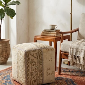 Pode incluir: Um pufe quadrado estofado em tecido entrelaçado bege e castanho com um padrão geométrico desbotado. O pufe tem uma forma estruturada e quadrada com costuras visíveis ao longo das bordas, exibindo um design têxtil texturizado e tonal.