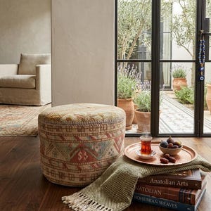 May include: A round, patterned ottoman in beige, red, and blue sits on a dark wood floor. A copper tray with a glass of tea and a bowl of figs rests on a stack of books and a green fringed throw. The scene is set near a window.