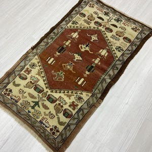 Puede incluir: Alfombra rectangular con un patrón central de rombos marrón rojizo con figuras y animales estilizados. La alfombra tiene un fondo beige con motivos geométricos y florales, enmarcado por un borde marrón oscuro. La alfombra mide aproximadamente 91 cm de largo y 61 cm de ancho.