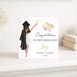 May include: A graduation card with an illustration of a graduate in a black gown and cap, holding a diploma and gold balloons. The card reads "Congratulations on your Graduation" with the name "Lucy" and "Teesside University 2026".
