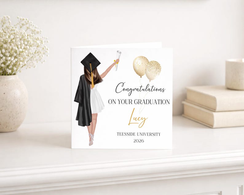 May include: A graduation card with an illustration of a graduate in a black gown and cap, holding a diploma and gold balloons. The card reads "Congratulations on your Graduation" with the name "Lucy" and "Teesside University 2026".