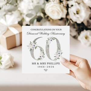 May include: A square white greeting card featuring the text CONGRATULATIONS ON YOUR Diamond Wedding Anniversary above a large silver glitter number 60 decorated with white floral accents. Below is MR & MRS PHILLIPS 1966-2026 and a diamond icon.