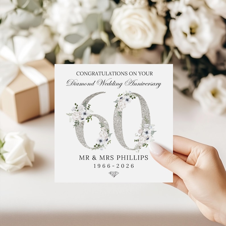 May include: A square white greeting card featuring the text CONGRATULATIONS ON YOUR Diamond Wedding Anniversary above a large silver glitter number 60 decorated with white floral accents. Below is MR & MRS PHILLIPS 1966-2026 and a diamond icon.