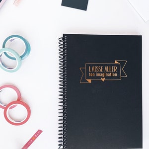 May include: A black spiral bound notebook with the words "LAISSE ALLER ton imagination" in gold lettering on the front cover. The notebook is surrounded by various colourful washi tapes and stickers.
