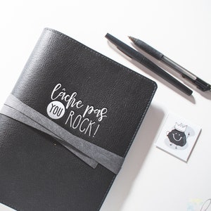 Peut inclure: Un carnet en cuir noir avec une fermeture en ruban gris. Le carnet porte l'inscription blanche "L&acirc;che pas you rock!"
