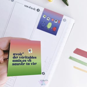 May include: A planner page with a colourful sticker featuring cartoon characters and a string of lights. A hand holds a square sticker with French text. Yellow and green pens, a pink paperclip, and decorative tape are also visible.