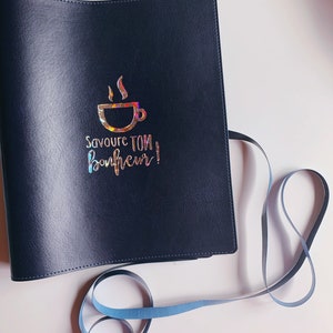 Peut inclure: Un carnet reli&eacute; en cuir noir avec un motif en feuille d'or d'une tasse de caf&eacute; et le texte "Savoure ton bonheur!"