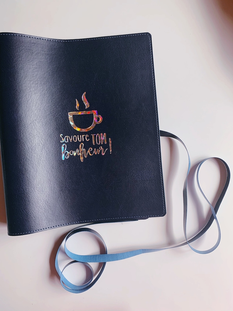 Peut inclure: Un carnet reli&eacute; en cuir noir avec un motif en feuille d'or d'une tasse de caf&eacute; et le texte "Savoure ton bonheur!"
