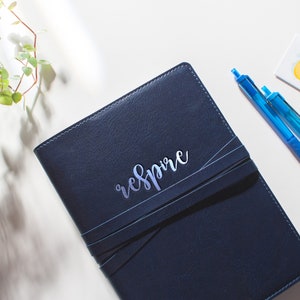 Peut inclure: Un journal reli&eacute; en cuir bleu fonc&eacute; avec une inscription argent&eacute;e qui dit "respire". Deux stylos bleus sont pos&eacute;s sur une surface blanche &agrave; c&ocirc;t&eacute; du journal.