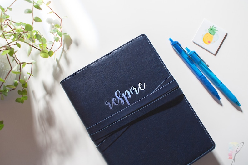 Peut inclure: Un journal reli&eacute; en cuir bleu fonc&eacute; avec une inscription argent&eacute;e qui dit "respire". Deux stylos bleus sont pos&eacute;s sur une surface blanche &agrave; c&ocirc;t&eacute; du journal.