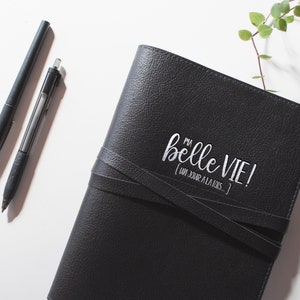 Peut inclure: Un journal en cuir noir avec une inscription blanche qui dit "Ma belle vie! (Un jour a la fois)". Le journal a une fermeture &agrave; boucle en cuir et est pos&eacute; sur une surface blanche.