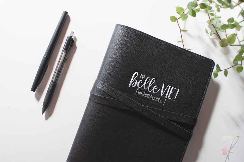 Peut inclure: Un journal en cuir noir avec une inscription blanche qui dit "Ma belle vie! (Un jour a la fois)". Le journal a une fermeture &agrave; boucle en cuir et est pos&eacute; sur une surface blanche.