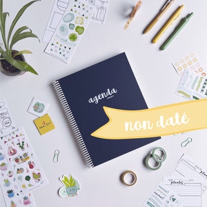 Peut inclure: Un agenda bleu avec le mot "agenda" sur la couverture. L'agenda est ouvert à une page avec les mots "non daté" écrits en jaune. L'agenda est entouré de divers autocollants, rubans adhésifs washi et stylos.