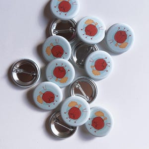 May include: A collection of ten round, white button badges featuring a cartoon beetroot design. Each beetroot has a red body, a green stem, and a frowning face. The badges have silver metal backs with safety pins.