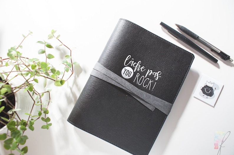 Peut inclure: Un carnet en cuir noir avec une fermeture en ruban gris. Le carnet porte l'inscription blanche "L&acirc;che pas you rock!"