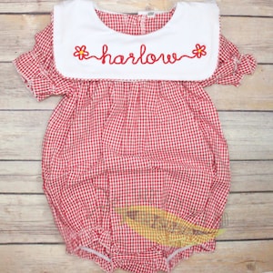 May include: Red and white gingham baby onesie with a white Peter Pan collar and the name "harlow" embroidered on the collar in white with yellow flowers on either side.