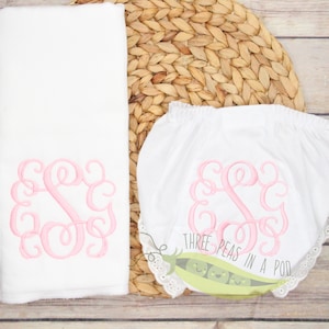 May include: A white baby diaper with pink embroidered monogram and lace trim. The diaper is on a woven straw mat. The text "Three Peas in a Pod" is on the diaper.