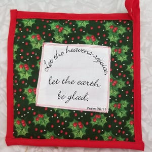 May include: Square, quilted hot pad with a red border and hanging loop. The center features a green holly and berry pattern, with a white square containing the text "Let the heavens rejoice, let the earth be glad. Psalm 96:11."