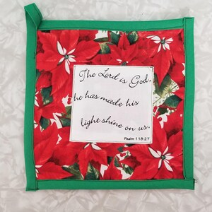 May include: Square Christmas potholder with a red poinsettia floral pattern and a green border. The center features a white rectangle with the text "The Lord is God, he has made his light shine on us. Psalm 118:27."