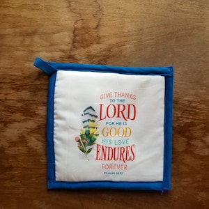 May include: White pot holder with blue trim and a colorful floral design. The text reads "Give thanks to the Lord for he is good. His love endures forever. Psalm 107:1."