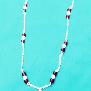 May include: A white beaded necklace with alternating red and clear beads. A small silver cross pendant hangs from the bottom. The necklace is displayed on a teal background.