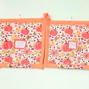 May include: Two square fabric potholders with orange borders and a pattern of pumpkins, autumn leaves, and apples. Each features a small rectangular patch with printed text, including the Bible verse Psalm 100:4 on one and Psalm 136:1 on the other.