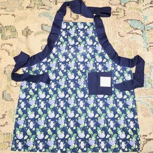 May include: A blue apron with a floral pattern of white and purple flowers. The apron has a dark blue trim and pocket. The ties are also dark blue. The apron is designed for kitchen or craft use.