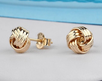 May include: A pair of gold-colored knot stud earrings. The earrings have a textured, woven design, and are displayed on a white surface with a blue background. The earrings are small and delicate, suitable for everyday wear.