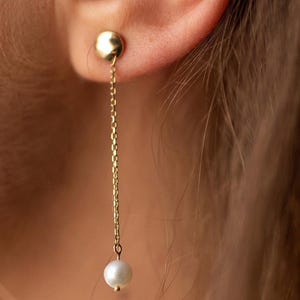 May include: A gold-colored earring with a small, round stud and a delicate chain. The chain suspends a white pearl. The earring is shown in close-up, highlighting the details of the jewelry.