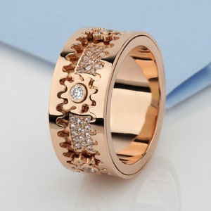 May include: A rose gold band ring featuring a mechanical gear-inspired design. The outer surface alternates between smooth rose gold sections and recessed panels encrusted with small clear stones, with a single larger stone set in the center.