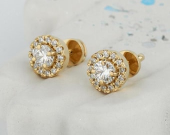 May include: A pair of gold stud earrings featuring a round, clear stone set within a halo of smaller stones.