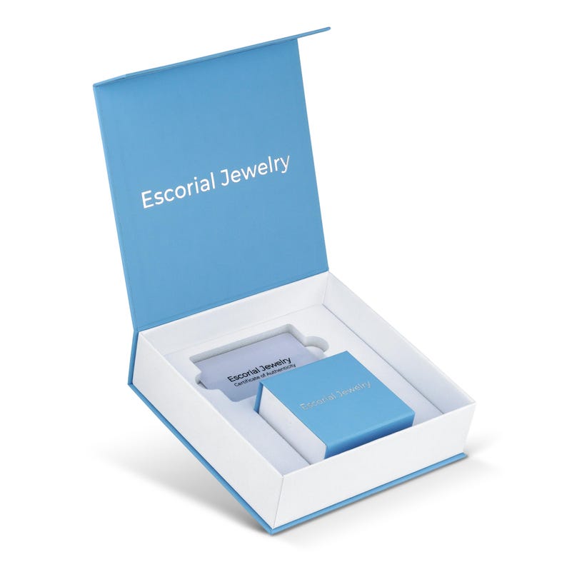 Op de afbeelding: Een lichtblauw sieradendoosje met een witte binnenkant, voorzien van de tekst Escorial Jewelry op het deksel. Binnenin bevinden zich een kleiner bijpassend blauw doosje en een rechthoekige kaart met de tekst Escorial Jewelry Certificate of Authenticity.