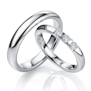 Op de afbeelding: Twee in elkaar grijpende ringen met een gepolijste zilverkleurige afwerking. De grotere ring heeft een gladde, effen band. De kleinere ring is bezet met drie ronde, heldere, gefacetteerde stenen in een rij, vastgezet met metalen pootjes.