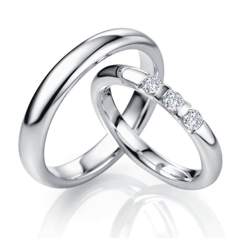 Op de afbeelding: Twee in elkaar grijpende ringen met een gepolijste zilverkleurige afwerking. De grotere ring heeft een gladde, effen band. De kleinere ring is bezet met drie ronde, heldere, gefacetteerde stenen in een rij, vastgezet met metalen pootjes.