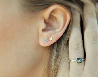 May include: A person wearing a pair of small square earrings in silver and gold. The person is also wearing a ring with a dark green gemstone.