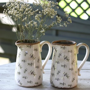 May include: Two off-white ceramic pitchers with a floral pattern and brown trim. One pitcher holds dried white flowers. The pitchers have curved handles and are displayed on a wooden surface. The pitchers are decorative.