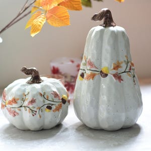 Op de afbeelding: Twee witte keramische pompoenen met een glanzende, gespikkelde afwerking en bruine stelen. De pompoenen zijn versierd met verhoogde, geschilderde motieven van herfstbladeren en kleine eikeltjes die rond het midden van elk stuk lopen.