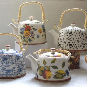 May include: Four ceramic teapots with bamboo handles and lids. One features blue floral patterns, one has colourful pansies, one displays a black floral print, and one shows a fruit motif. Two small ceramic chicken figurines are positioned nearby.