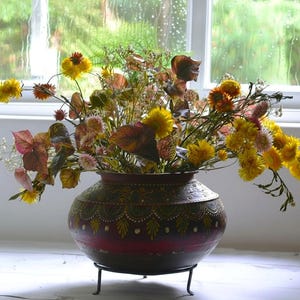 Peut inclure: Un pot en métal rond de couleur sombre avec une large ouverture et des motifs décoratifs peints à la main en jaune et vert, reposant sur un support tripode en métal noir. Le pot est rempli d'un arrangement de fleurs séchées jaunes et roses.