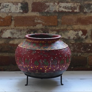 Peut inclure: Un pot en terre cuite rouge de forme arrondie, orné de motifs floraux peints à la main en vert et jaune ainsi que de points blancs. Le pot présente une large ouverture et repose sur un support tripode en métal noir, avec une texture usée.