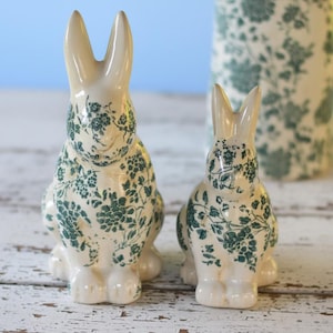 Op de afbeelding: Twee keramische konijnenbeeldjes met een crèmekleurige basis en een groen bloemmotief. De konijnen zitten met rechte oren. Op de achtergrond is een bijpassende vaas te zien. De objecten staan op een wit houten oppervlak.