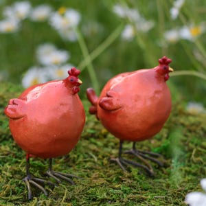May include: Two round, red ceramic chicken figurines with smooth, glossy surfaces, small red combs, and thin, dark metal wire legs. Each bird features a plump, spherical body and a small, pointed beak, standing upright on a textured green surface.