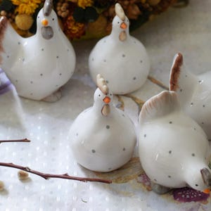 May include: Five white ceramic hens with black polka dots and orange beaks. The hens are arranged on a white surface with a floral pattern.