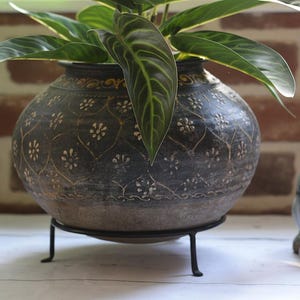 May include: A round, dark grey ceramic planter with a weathered finish, featuring a repeating gold-toned floral pattern and a scalloped grid design. The planter rests on a black metal wire stand and contains a green plant with veined leaves.